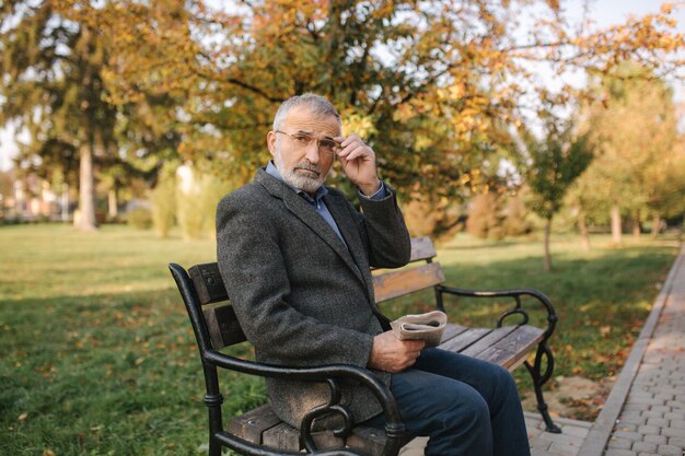 Bearded elderly man in glasses reading newspaper in the autumn park Handsome grayhaired man sitting on the bench early in the morning