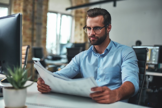 Bearded engineer in eyeglasses and formal wear looking at blueprint while sitting in the office