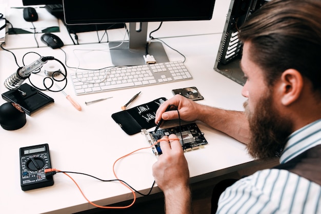 ひげを生やしたエンジニアがマイクロサーキットをテストしています。修理工場で電子機器を修理するエンジニアの職場