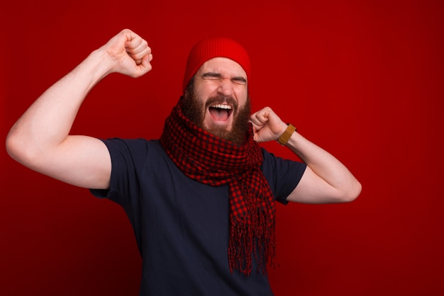 Bearded excited man with a scarf and a hat on, is screaming of joy near red wall.