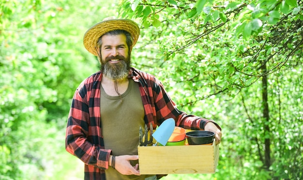 Bearded farmer in straw hat with wooden box mature man gardener working in greenhouse brutal male carry garden tools human and nature farming and floral concept spring season