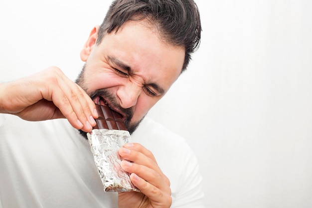 Bearded fat man biting chocolate. it is hard for him to do that. Young man holds bar with both hands.