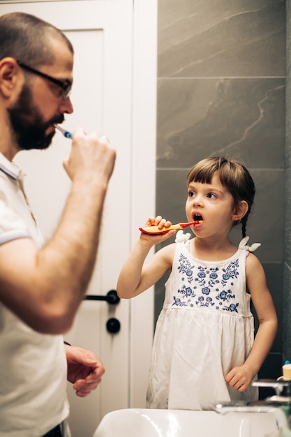 Padre barbuto e la sua piccola figlia che si lavano i denti in un bagno e si guardano l'un l'altro. uomo che insegna a sua figlia come lavarsi i denti.