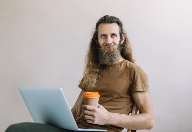 Libero professionista barbuto usando il portatile, bere caffè, lavorando da casa