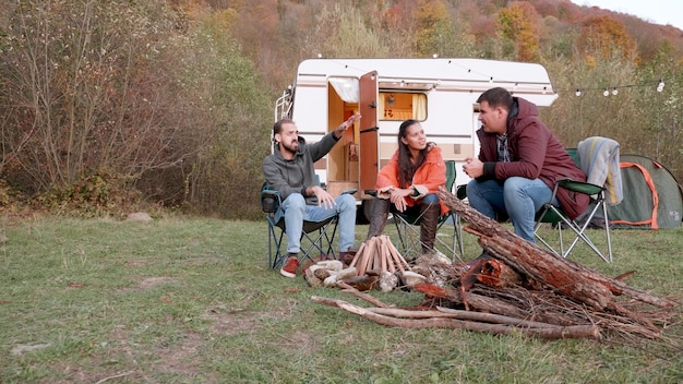Bearded friend telling a story to his couple friends in front\
of camper van. wood for fire. friends relaxing.