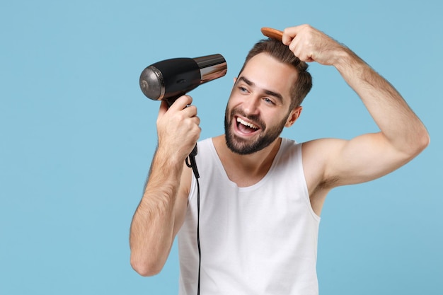 Bearded fun young man 20s years old in white shirt hold blow dry hair isolated on blue pastel wall background, studio portrait. Skin care healthcare cosmetic procedures concept. Mock up copy space.