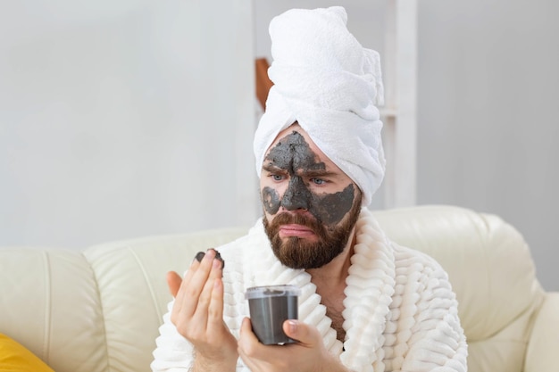 Bearded funny man having fun with a cosmetic mask on his face made from black clay men skin care