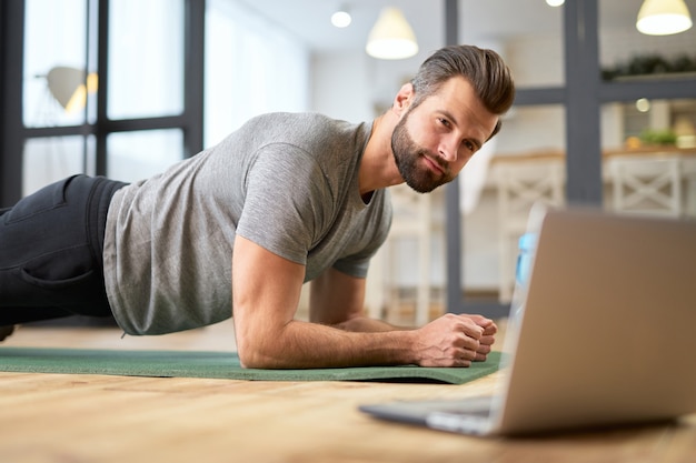 Bearded gentleman watching online workout on notebook and doing exercise at home