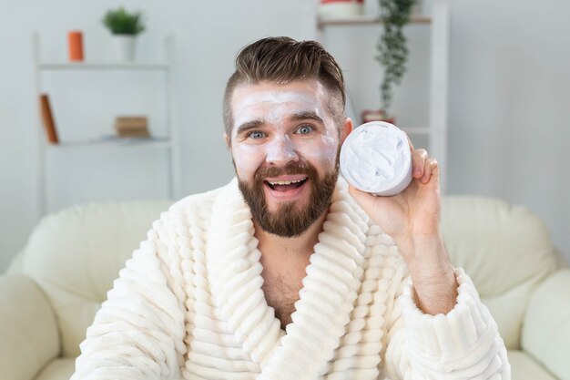 Ragazzo barbuto che applica la crema per il viso davanti allo specchio per la cura della pelle e la spa per il concetto dell'uomo