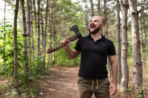 Bearded guy in a black Tshirt stands in a summer forest with an ax in his hand and smiles Lumberjack posing for the camera Chopping wood Natural landscape