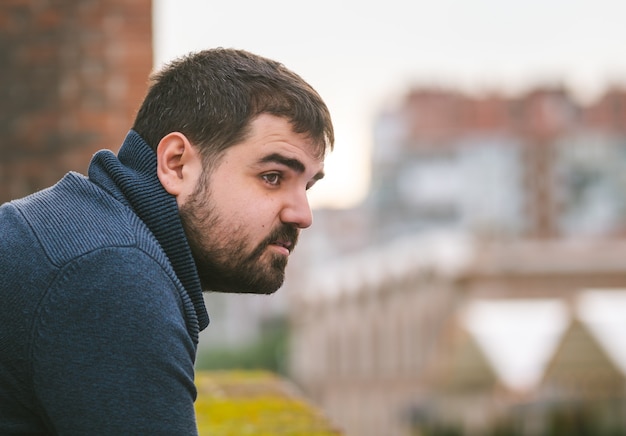 Bearded guy in dark blue sweater leaning on a railing on the street looking aside seriously