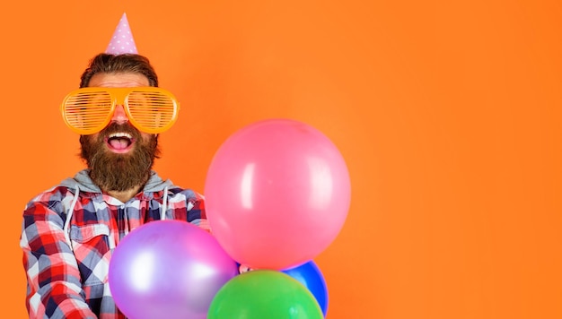 Foto ragazzo barbuto con occhiali divertenti e cappello a cono con palloncini uomo sulla festa di buon compleanno che celebra