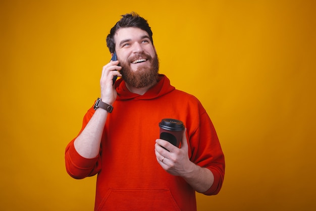 ひげを生やした男が黄色いスペースでテイクアウトのコーヒーを楽しみながら電話で話している。