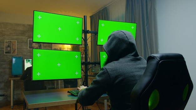 Bearded hacker hiding his face wearing a hoodie using computer with green screens.