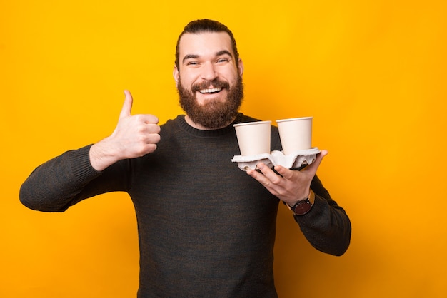 Bearded handsome man showing thumb up and holding take away coffee over yellow