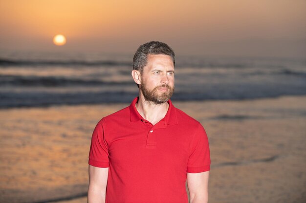 Bearded handsome man in summer shirt on beach with sunset over sea, relax.