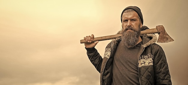 Bearded handsome serious man with rusty axe against cloudy sky