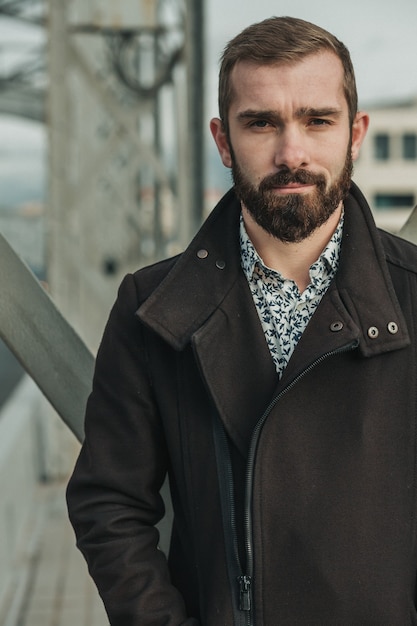 Bearded handsome young man in front of bridge construction with flirtation and slight smile. He is wearing brown jacket and light shirt.