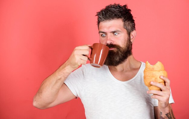 Bearded hipster enjoy breakfast drink coffee Morning tradition concept Unhealthy but yummy breakfast Fresh baked croissant Delicious breakfast Man start morning with coffee and croissant