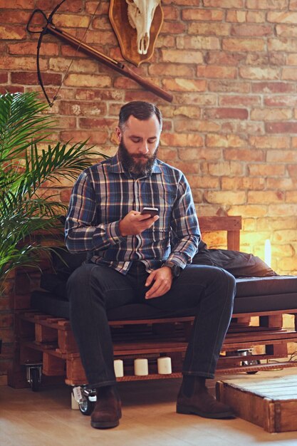 A bearded hipster male dressed in a fleece shirt using smartphone in a room with loft interior.