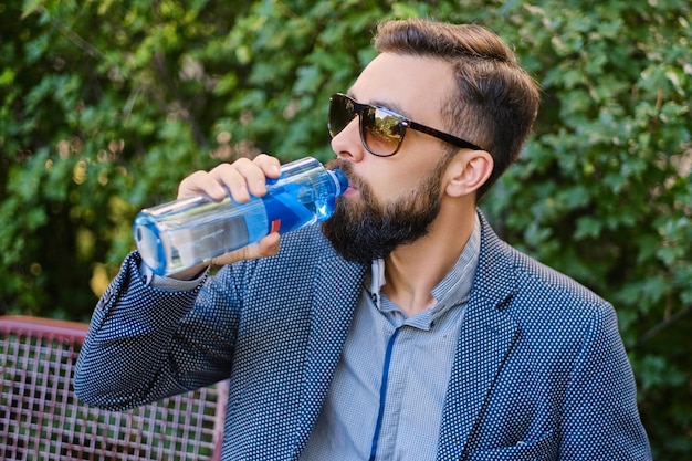 Bearded hipster male in sunglasses, drinks mineral water.