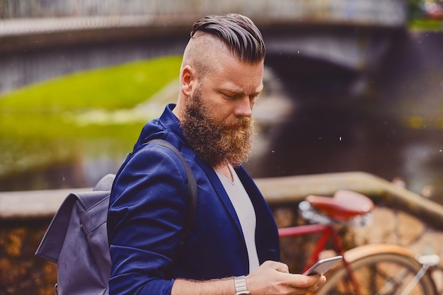 川の近くの公園でスマートフォンを使用してひげを生やしたヒップスターの男性。