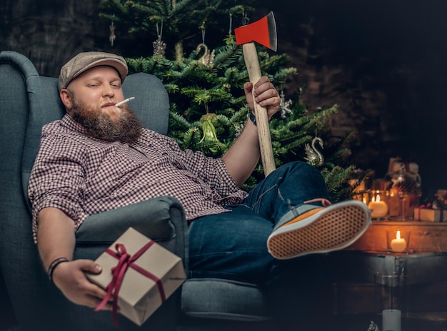 Bearded hipster man sits on a chair and holds an axe over Christmas background with fir tree.