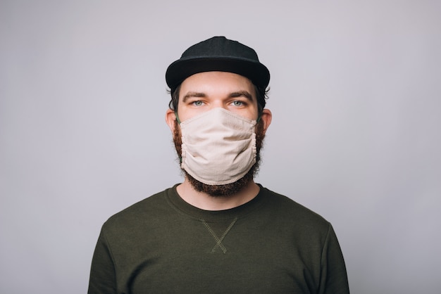 Bearded hipster man standing over white wall and wearing facial mask for virus