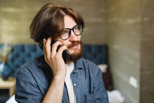 Foto uomo barbuto hipster con gli occhiali che parla sullo smartphone in camera da letto