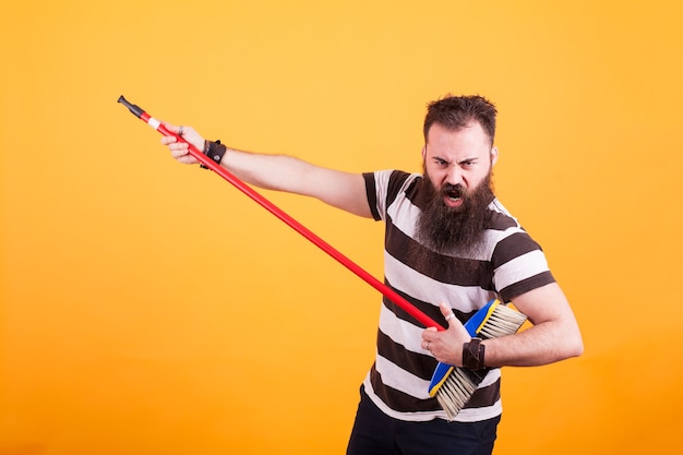 Bearded hipster in striped t-shirt using broom as if was a guitar over yellow background. Cool man.,