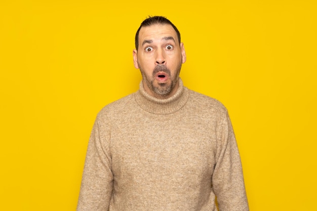 Bearded hispanic man in his 40s wearing a beige turtleneck tremendously amazed and surprised with open mouth in shock isolated over yellow background