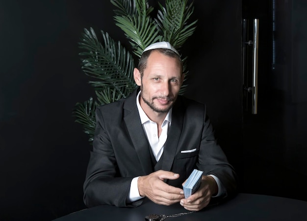 Bearded Jewish man in Yarmulke national hat Kippah sit at desk looking at camera holding hundred dollar bills Jew Israeli businessman with cunning eyes in office peeks out from under a table