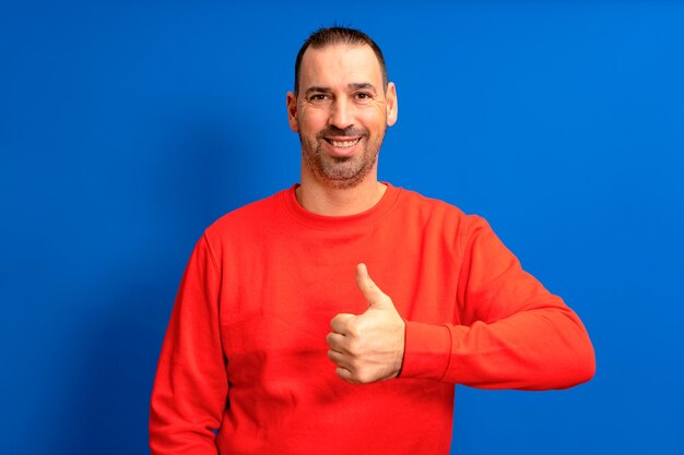 Bearded latin man wearing a red sweater raising his thumb up in approval while smiling at the camera in a friendly attitude isolated over blue background person