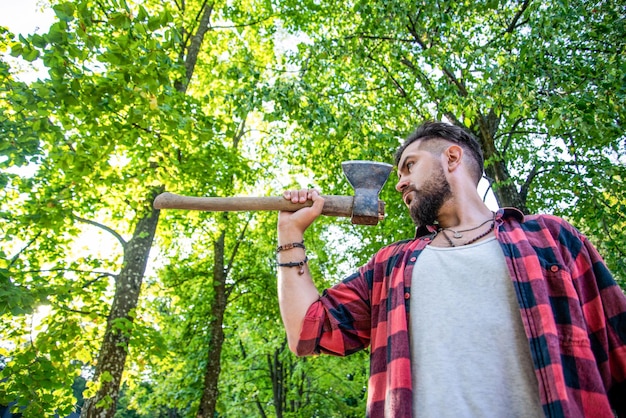 Bearded lumberjack with a large ax bearded guy in a summer\
forest with an ax in his hand lumberjack man chopping wood brutal\
lumberjack bearded man in woods on a background of trees