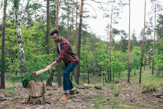 Bearded lumberman in plaid shirt and denim jeans cutting wood with ax in forest