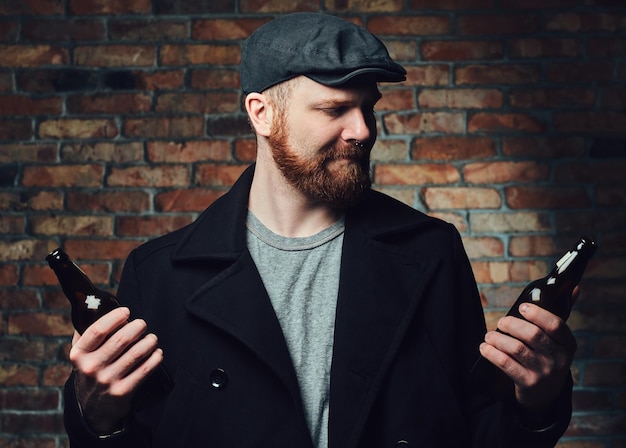 Bearded male dressed in a black tweed flat cap and wool jacket holds beer bottle over the wall of a brick.