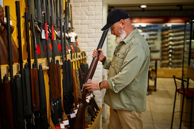 銃の店で狩猟用ライフルの列を持つショーケースでひげを生やした男性ハンター。武器屋のインテリア、弾薬と弾薬の品揃え、銃器の選択、射撃の趣味とライフスタイル