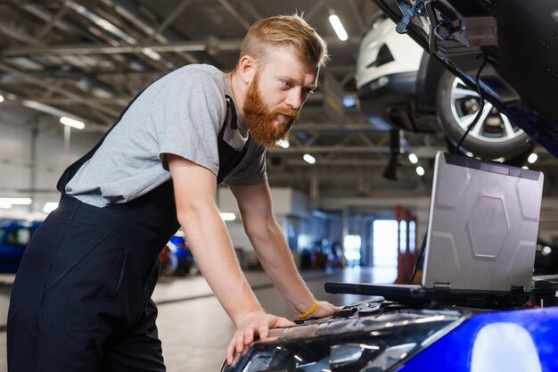 あごひげを生やした男性の機械技師が、サービス ステーションで車のシステムをチェックしながら、診断用ラップトップを車に取り付けました。