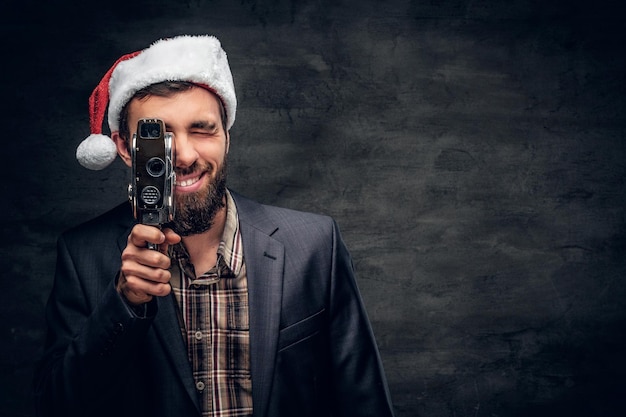 Bearded male in Santa's hat holds the old 8mm video camera on grey vignette background.