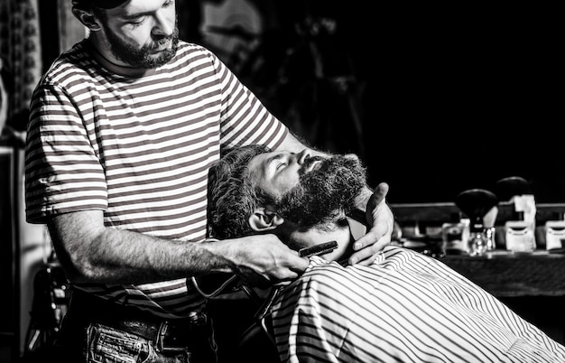 Foto uomo barbuto seduto su una poltrona in un barbiere mentre il parrucchiere si rade la barba con un pericoloso rasoio. barbiere che rade un uomo barbuto in un negozio di barbiere. bianco e nero.