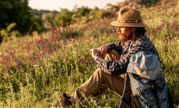 Bearded male with backpack sitting on grass during trip