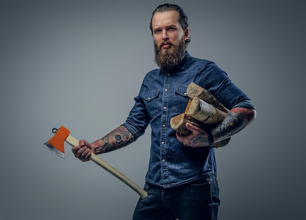 Bearded male with tattoos on his arms holds an axe and firewoods.