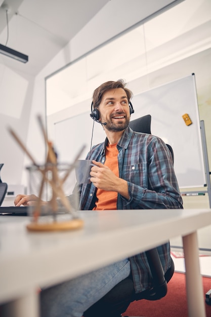 Bearded male worker in headset holding cup of coffee and smiling while sitting at the table with computer