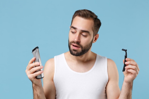 Bearded man 20s years old in white shirt hold choice electric\
plastic razor isolated on blue pastel background studio portrait.\
skin care healthcare cosmetic procedures concept. mock up copy\
space.