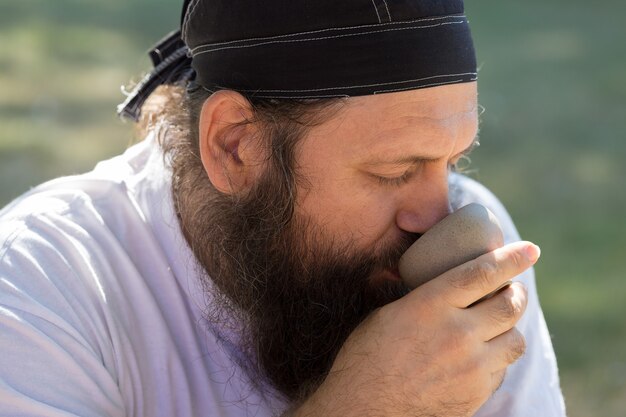 バンダナのひげを生やした男が自然を背景にお茶を飲んでいます。