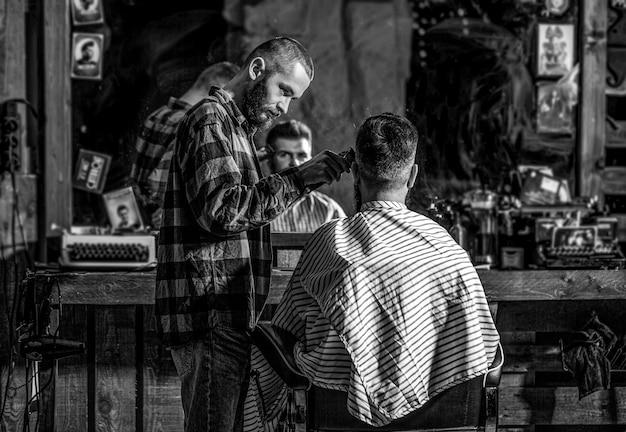 Бородатый мужчина в парикмахерской Концепция стрижки Руки парикмахера с машинкой для стрижки волос Черно-белый