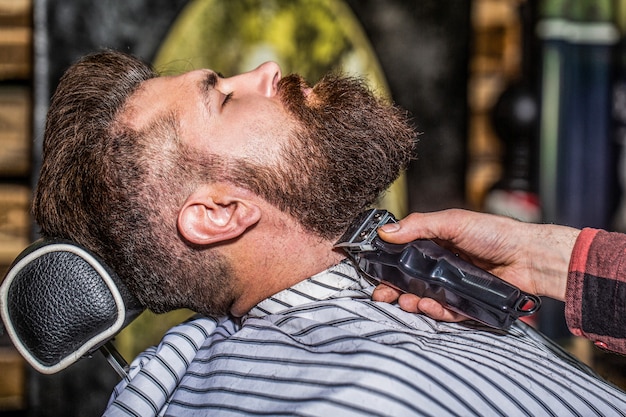 Bearded man in barbershop. Man visiting hairstylist in barbershop. Barber works with a beard clipper. Hipster client getting haircut. Hands of a hairdresser with a beard clipper, closeup.