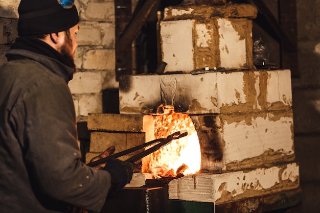 Bearded man blacksmith gets a red-hot billet out of the furnace.