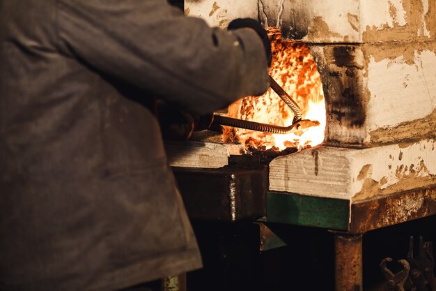 Bearded man blacksmith gets a red-hot billet out of the furnace.