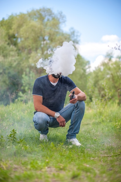 Бородатый мужчина дует много дыма, используя электронную сигарету vape на лесной земле